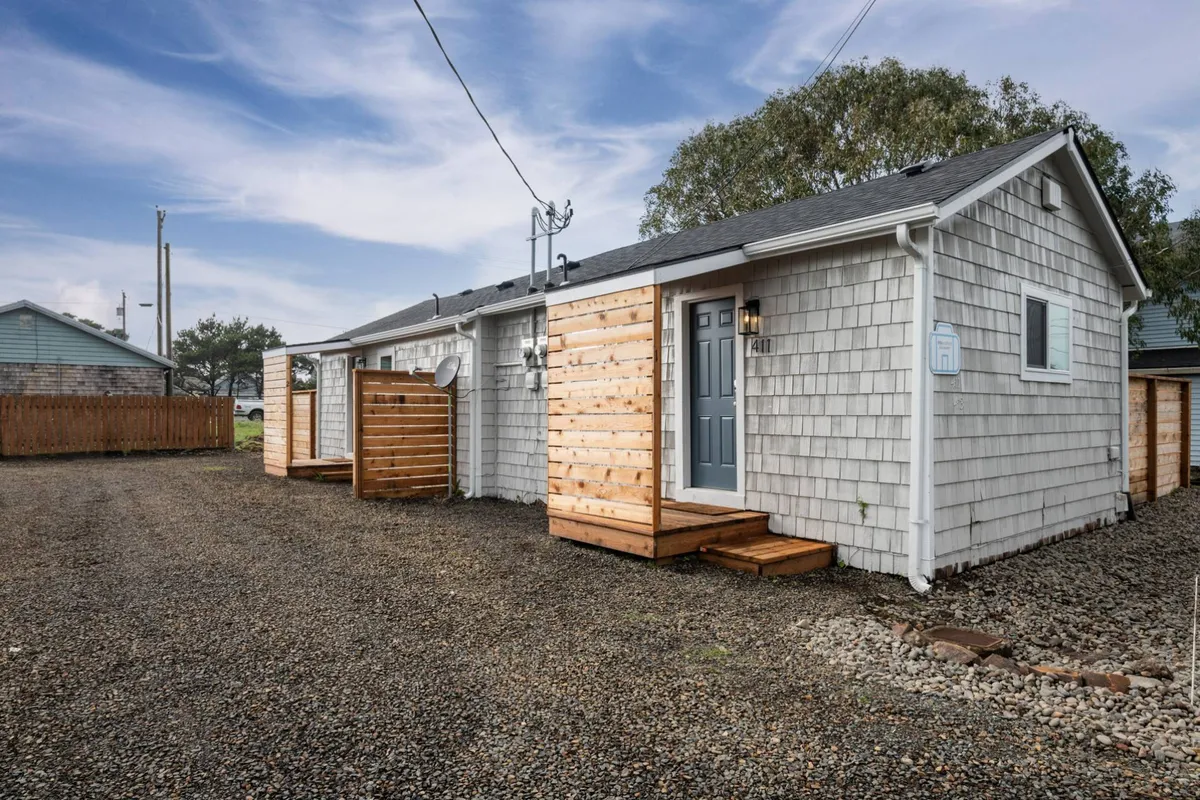 This image features the welcoming front porch of Coral Cottage Unit 1, with gray shingle siding, wooden steps, and a decorative light by the door. The setting hints at the cottage's inviting atmosphere and its proximity to neighborhood amenities, suggesting ease of coming and going for local adventures.