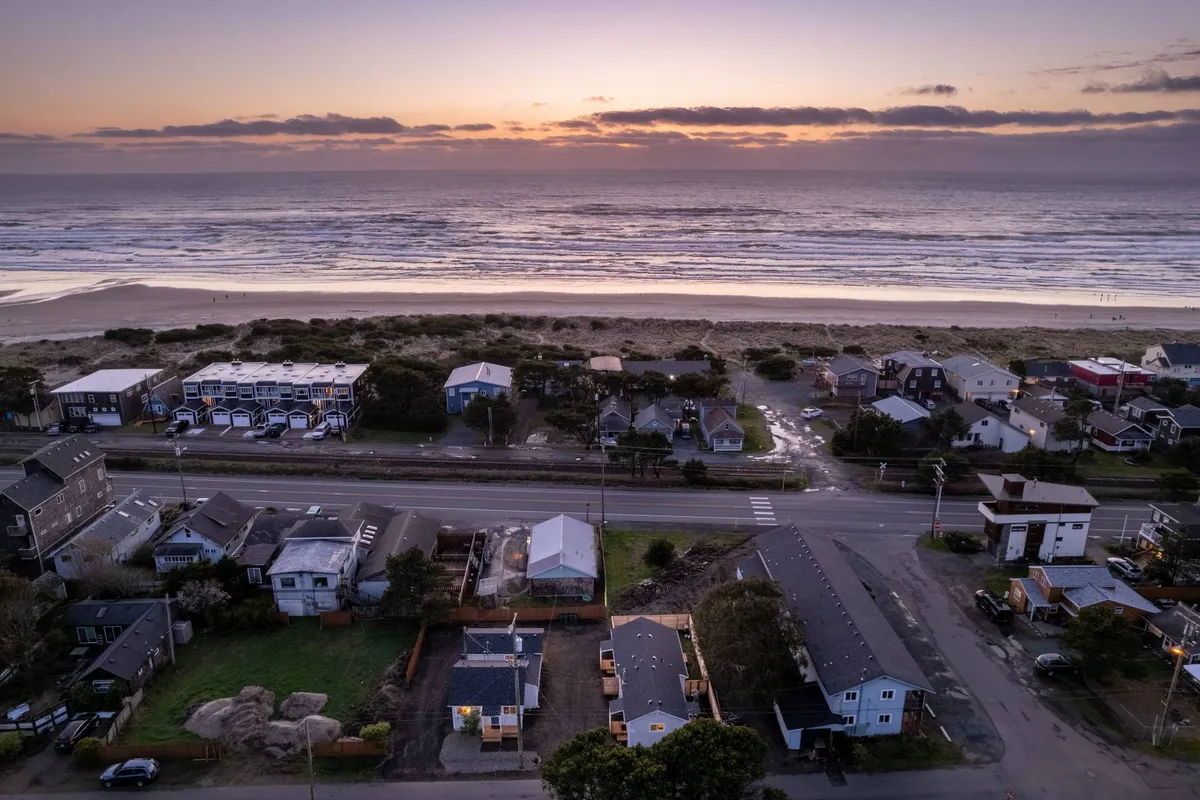 The photograph features calm ocean waves rolling onto shore at sunset, with houses lining the water’s edge and a glowing sky overhead. The tranquil mood and visible community connection highlight evening strolls, sunset-viewing, and the allure of a walkable, coastal neighborhood.