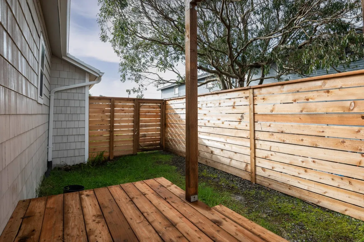 A peaceful exterior view featuring a wooden deck bordered by a fenced grassy area, shaded by a mature eucalyptus tree. The space invites outdoor relaxation and provides an open environment ideal for pets to explore safely. The warm sunlight on the deck and greenery evoke a tranquil, pet-friendly atmosphere.