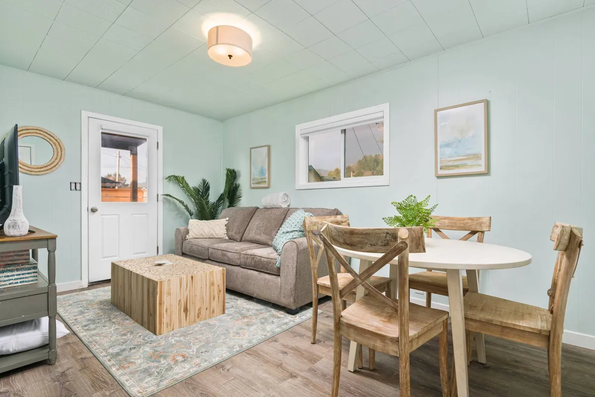 The image showcases a bright, tastefully decorated living and dining area with a plush sofa, inviting dining table, lush decorative plant, and cheerful coastal artwork on the walls. The space feels open yet cozy, ideal for relaxing, sharing meals, and spending quality time together.