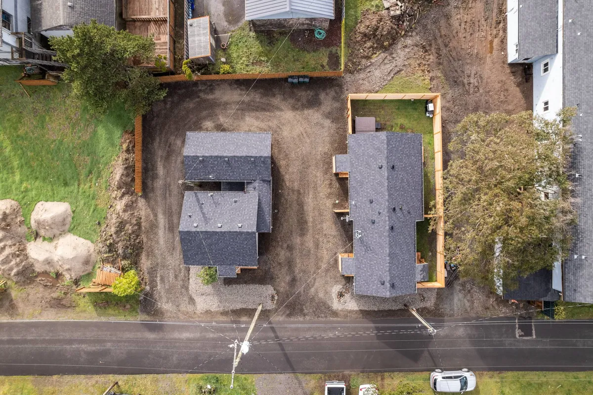 A wide exterior view featuring two adjacent houses with a gravel driveway and a lush green yard in the foreground. The homes are set in a friendly, open space suitable for gatherings or play, offering a sense of arrival and neighborhood vibe.