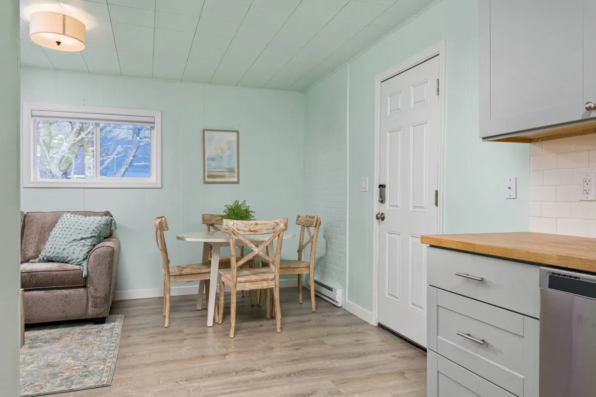 This image captures the heart of the open-concept living space, featuring a comfortable sofa, a dining table, a kitchen counter, and a large window filling the room with natural light. The layout showcases both relaxation and dining areas seamlessly integrated, while sunlight streams through, emphasizing the welcoming and comfortable atmosphere.
