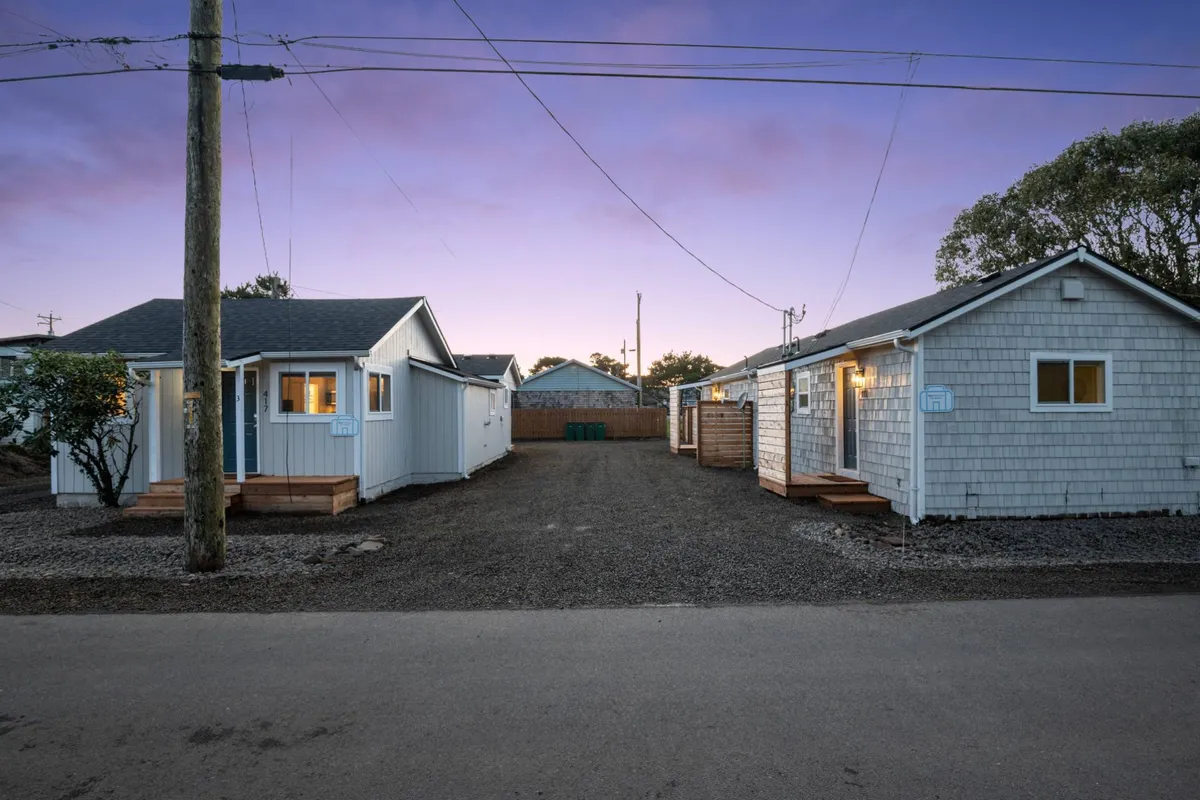 Two charming neighboring cottages are visible along a gravel driveway, each with welcoming wooden decks. The setup highlights the small complex’s sense of community as well as the privacy of each unit, with plenty of outdoor space for pets and socializing with neighbors.