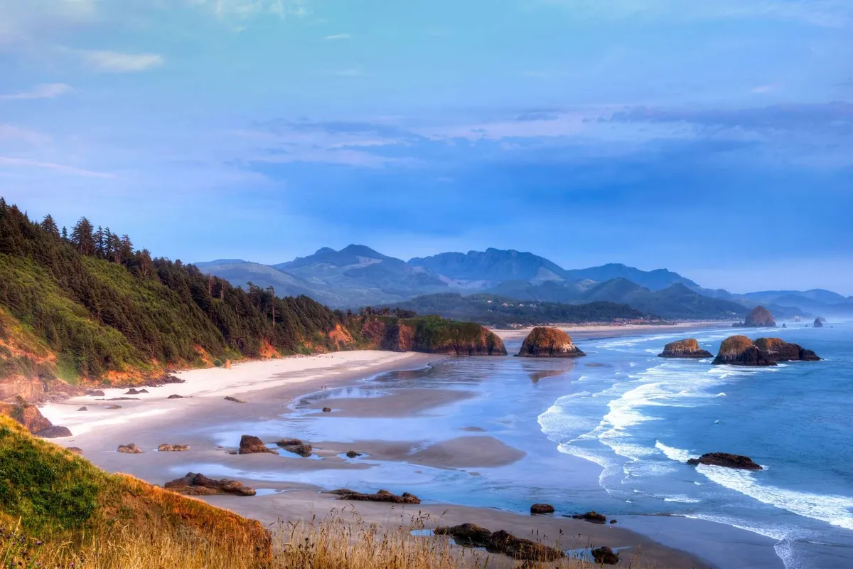 A sweeping, panoramic view displays the rugged expanse of Rockaway Beach, with ocean waves, dramatic rock formations, and forested cliffs leading to distant mountains. The image embodies the spirit of adventure and the natural beauty that defines the area, suggesting opportunities for exploring, beachcombing, and sunset viewing.